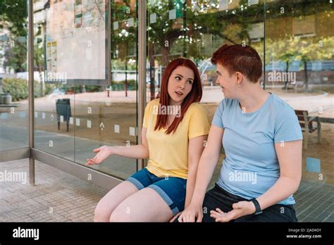 Two Happy Young Friends Sitting At The Bus Stop Talking And Gossiping