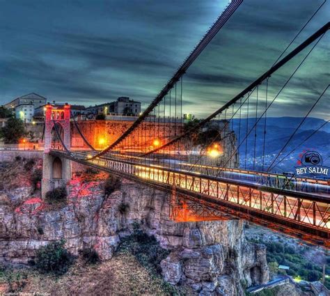 A Long Bridge With Lights On It Over A Cliff And Below Is A City At Night