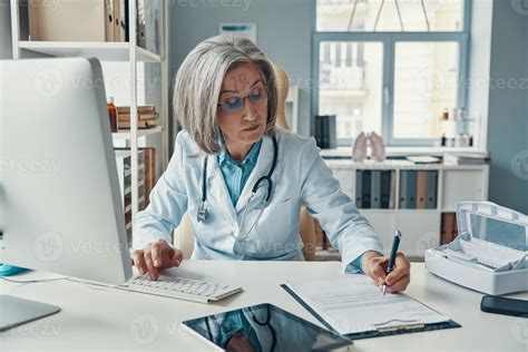 Serious Mature Female Doctor In White Lab Coat Writing Something Down
