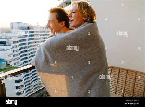 European Gay Couple Wrapped Blanket Laughing And Hugging On Balcony At