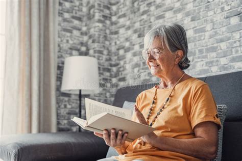 Happy Older Blonde Woman Sitting On Comfortable Sofa In Living Room Reading Interesting Book