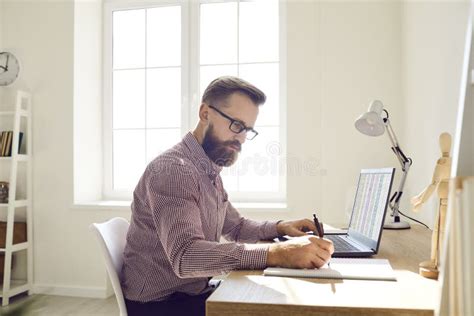 Businessman In Glasses Sitting At Desk Working On Laptop Computer And Taking Notes Stock Image