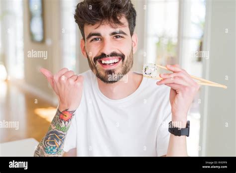 Young Man Eating Asian Sushi Using Chopsticks Pointing And Showing With Thumb Up To The Side