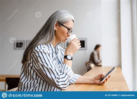 Focused White Haired Mature Woman Drinking Coffee And Using Cellphone