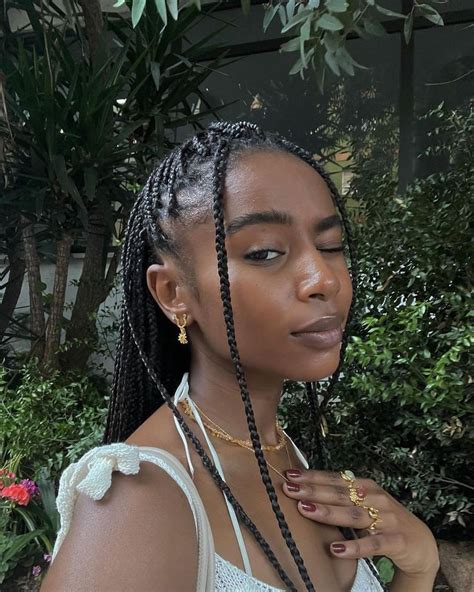 A Woman With Long Braids Standing In Front Of Some Bushes And Trees