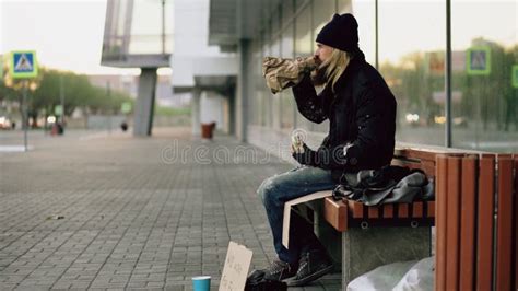 Homeless Young Man Eating Sandwich And Drinking Alcohol From Paper Bag