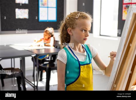 Caucasian Girl With Blonde Hair Paints On An Easel Wearing A Blue Apron In School Stock Photo