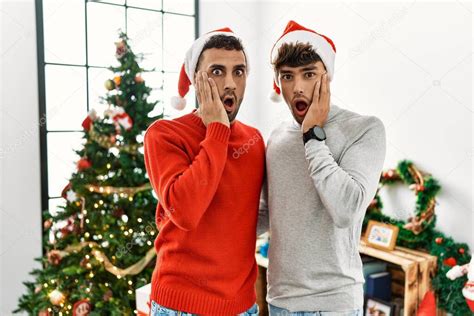 Joven pareja gay de pie junto al árbol de navidad vistiendo sombrero asustado y sorprendido