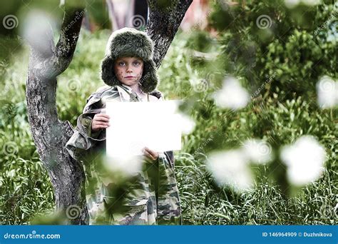 Boy In A Camouflage Uniform And A Hat With Earflaps Stands In An Apple