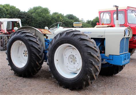 Two Large Tractors Parked Next To Each Other On A Dirt Field With Trees