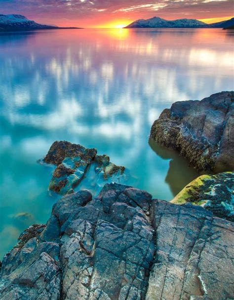 The Sun Is Setting Over Some Rocks In The Water And There Are Mountains