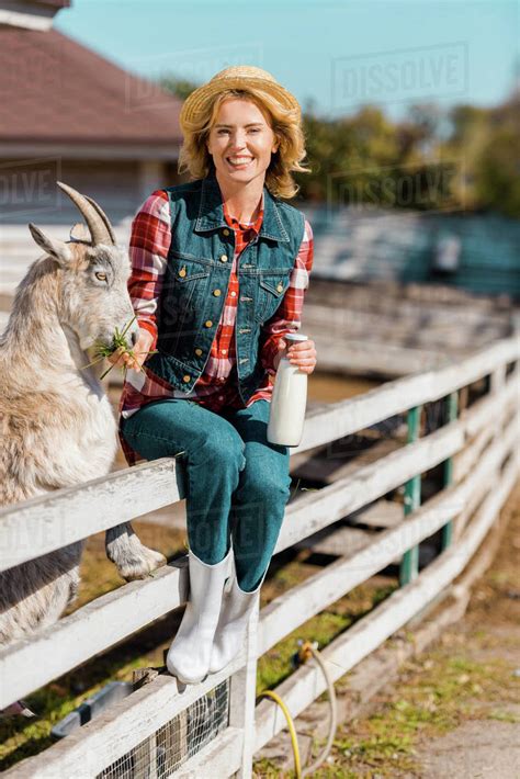 Smiling Female Farmer With Milk Bottle Sitting On Wooden Fence And