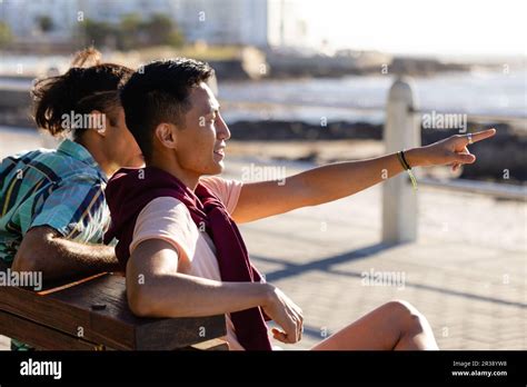 Happy Biracial Gay Male Couple Sitting On Bench And Pointing Away On Promenade By The Sea Stock