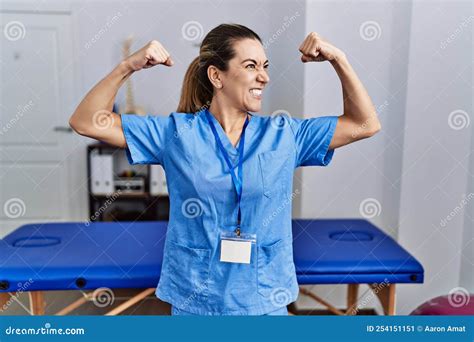 Young Hispanic Woman Wearing Physiotherapist Uniform Standing At Clinic Showing Arms Muscles