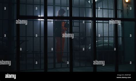 Prisoner in orange uniform kneels near bed, prays to God in prison cell holding Bible. Jailer ...