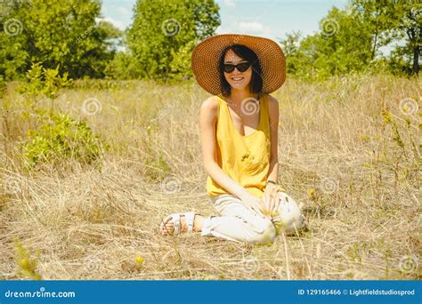 Attractive Woman In Sunglasses And Straw Hat Sitting On Dried Grass