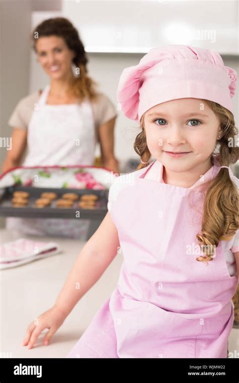 Cute Girl Wearing Pink Apron And Chefs Hat Smiling At Camera At Home In