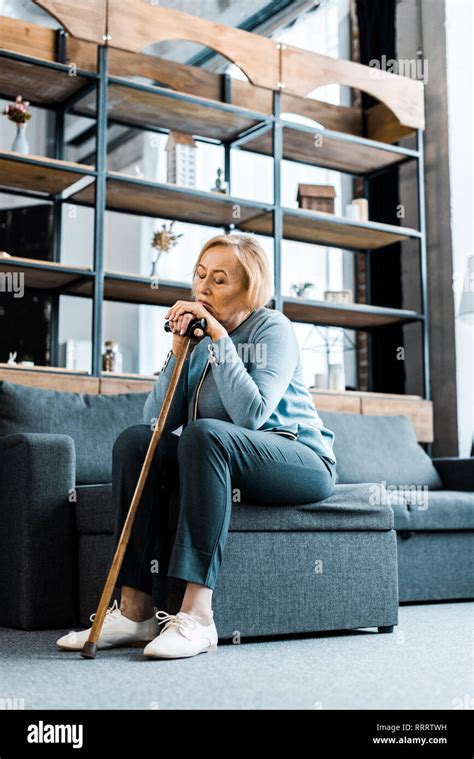 Sad Senior Woman Sitting On Couch And Leaning On Walking Stick In