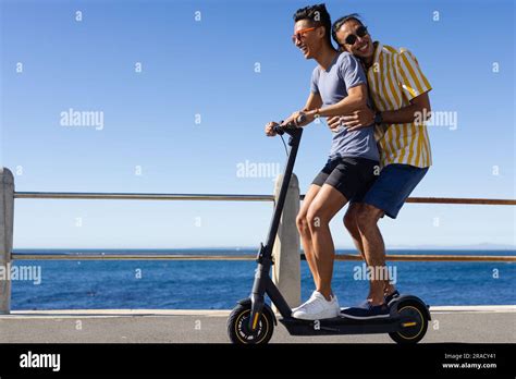 Happy Biracial Gay Male Couple Riding Scooter Together On Promenade By