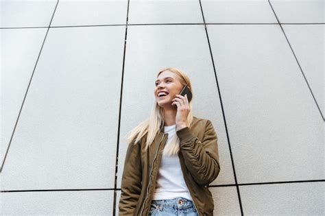 Free Photo Smiling Blonde Talking On Phone While Standing Near Wall