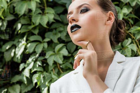 Elegant Young Woman In White Jacket Touching Face With Green Leaves At