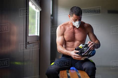 Strong Mature Male Firefighter With Naked Torso Sitting On Bench In Mask And Holding A Helmet