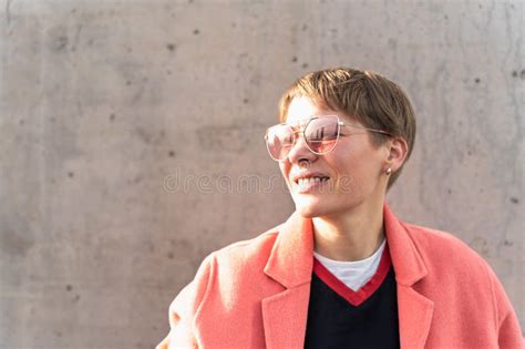 Modern Mature Woman With Glasses Wearing A Pink Jacket Smiling Against A Concrete Wall