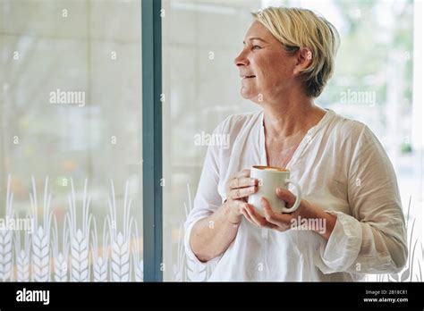 Positive Mature Woman In White Cotton Blouse Drinking Mug Of Coffee And