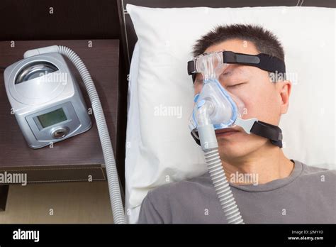 Asian man with sleep apnea using CPAP machine, wearing headgear mask connecting to air tube ...