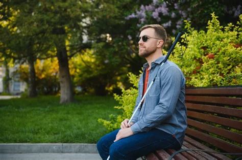 Visually Impaired Man With Walking Stick Sitting On Bench In City Park