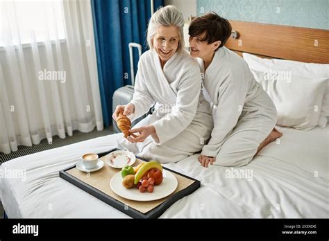 Senior Lesbian Couple Enjoying A Tender Moment Sitting On A Cozy Bed In A Hotel Room Stock