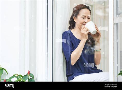 Portrait Of Smiling Mature Woman Sitting On Window Sill And Enjoying