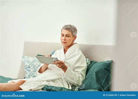 Smiling Mature Woman In Bathrobe Relaxing On A Bed Holding A Digital Tablet Stock Image Image