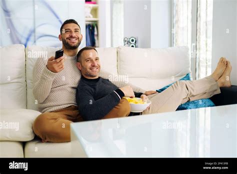 Gay Couple Sitting On The Couch At Home Watching Something On TV And Having A Snack Homosexual