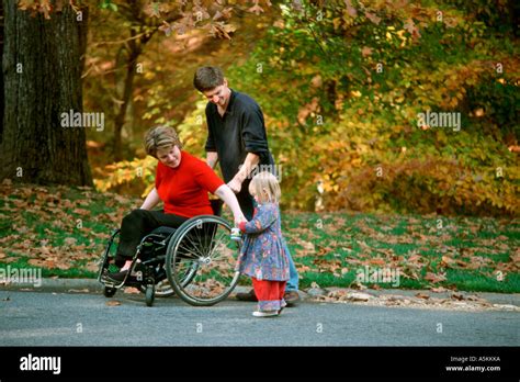 A Man Pushes His Wife In A Wheelchair Through A Park With Their Young Daughter Along Side Stock