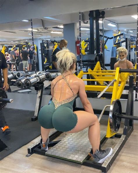 A Woman Squatting On A Bench In A Gym With Other People Working Out