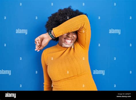 Black Woman With Curly Hair Standing Over Blue Background Covering Eyes With Arm Smiling