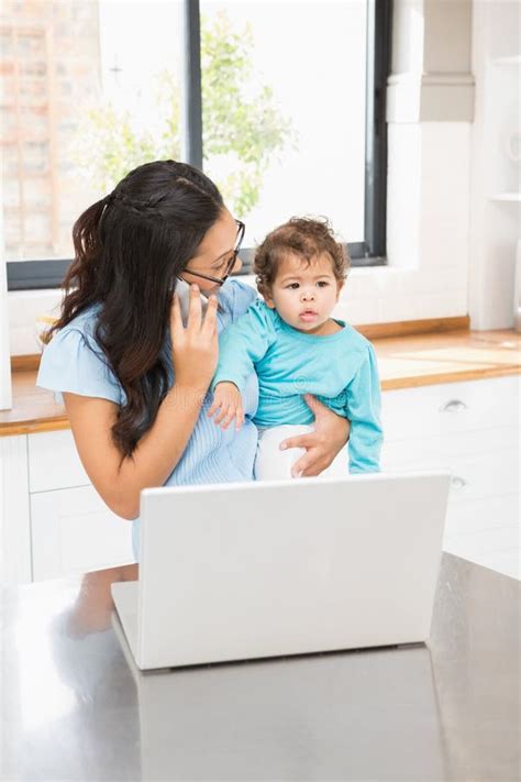 Smiling Brunette Holding Her Baby And Using Laptop On Phone Call Stock