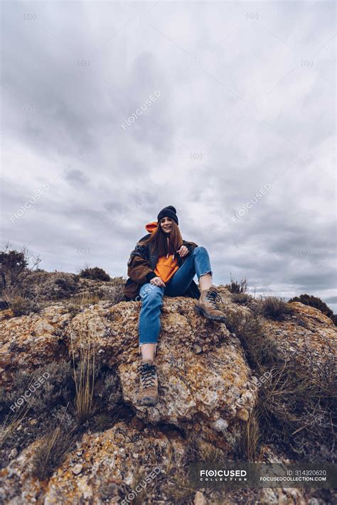 From Below Positive Woman Looking In Camera And Sitting On Rocks At