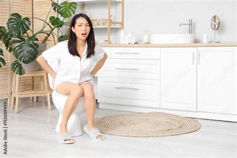 Young Asian Woman With Hemorrhoids Sitting On Toilet Bowl In Restroom