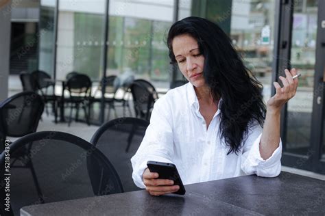 Mature Beautiful Woman Sitting At A Street Cafeteria With Smartphone And Checking Her Social