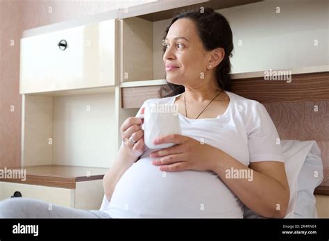 Authentic Adult Pregnant Woman With Cup Of Hot Tea Dreamily Looking Away Experiencing Happy