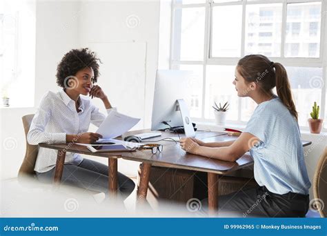 Two Young Women Talking Across Their Desks In An Office Stock Image