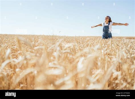 Delighted Female In Denim Overalls Standing In Wheat Field With