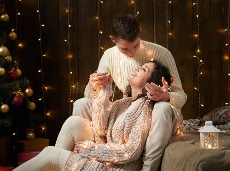 Young Couple Together In Christmas Lights And Decoration Dressed In White Fir Tree On Dark