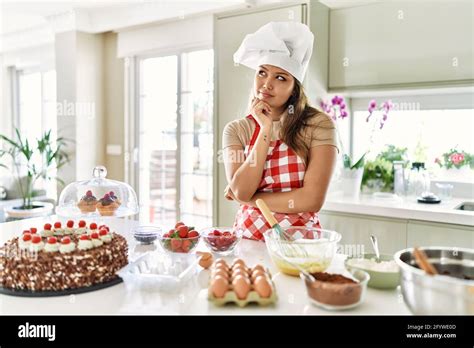 Beautiful Young Brunette Pastry Chef Woman Cooking Pastries At The Kitchen With Hand On Chin