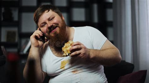 Dirty Fat Bearded Man Talks On The Smartphone Eating A Burger And