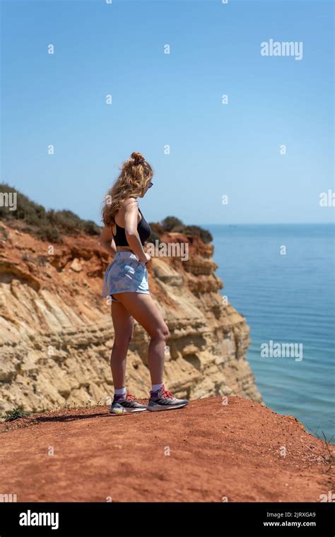 A young, blond woman posing on a cliff edge looking out over the ocean under the sun Stock Photo ...