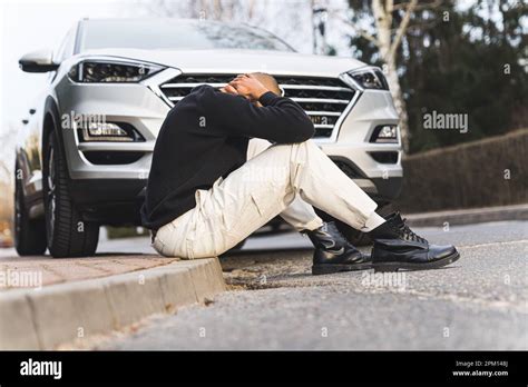 A Man Sitting On A Curb In Front Of A Grey Car With His Head Down No