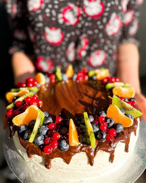 A Woman Holding A Cake With Fruit On Top And Chocolate Icing Drizzled